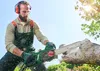 A man is using a chainsaw to cut a log outdoors on a sunny day. Produktbild-Vorschau