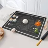 A black silicone baking mat with circular guides and rulers is laid out on a kitchen counter. It features a ball of dough, some steamed buns, shredded carrots, and broccoli florets, suggesting its use in baking and food preparation. A whisk, eggs, a wooden Voir les images du produit