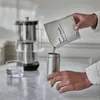 A person is pouring a powder from a pouch into a metal container, with a water filter pitcher and a glass of water in the background. Anteprima dell’immagine prodotto