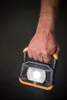 A hand holding a black and orange LED work light. Produktbild-Vorschau