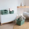 A green storage basket with a stuffed toy inside, placed on a jute rug next to a white dresser and a wooden desk. Anteprima dell’immagine prodotto