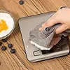 A hand is wiping a digital kitchen scale with a gray cloth, on a wooden table. There are blueberries and a bowl of flour with egg yolk nearby. Náhľad obrázku produktu