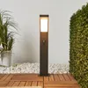 A modern, dark gray LED bollard lamp with a motion sensor is shown in an outdoor setting, placed on a wooden deck surrounded by white pebbles and lush green foliage. Produktbild-Vorschau