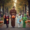A family is trick-or-treating on Halloween, walking down a tree-lined street with pumpkins and decorations. Náhled obrázku produktu