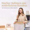 A woman is holding a beige laundry basket with a bamboo frame in a room with shelves and decorative items. Produktbild-Vorschau
