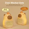Two cute, cartoon-like alarm clocks shaped like capybaras are displayed against a light brown background. Each clock has a digital display showing a different time and a speech bubble with text in German. Produktbild-Vorschau