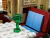 A close-up view of a Lego set, showing a green wine glass on a white surface with Lego studs, and a red and blue sofa-like structure in the background. Podgląd zdjęcia produktu