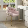 A pink armchair with gold legs is placed next to a white vanity table with drawers, in front of a window with a view of trees and a modern building. Produktbild-Vorschau
