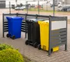 Three colored trash bins (blue, black, yellow) are placed in a metal enclosure with horizontal slats, on a paved surface. A green bush is in the foreground. Náhled obrázku produktu
