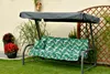 A garden swing with a patterned cushion and cover, set on a lawn next to plants and a fence. Produktbild-Vorschau