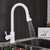 A white kitchen faucet with a pull-out spray head is shown in use, with water flowing into a dark sink. A framed picture and some decorative plants are visible in the background. Náhľad obrázku produktu