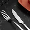 A close-up shot of a fork and knife on a black marble surface, with a portion of an orange plate in the background. Produktbild-Vorschau