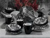 A collection of black and white patterned dinnerware, including plates, bowls, and mugs, arranged on a textured surface with rocks and foliage in the background. Náhľad obrázku produktu