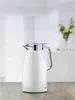 A white alfi thermos jug with a silver handle and lid sits on a wooden table. The background is blurred and shows a bright, airy room with some yellow flowers in a vase. Anteprima dell’immagine prodotto