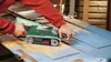 A person is using a Bosch belt sander to remove blue paint from a wooden door. Podgląd zdjęcia produktu
