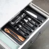 A black cutlery organizer filled with silverware and wooden utensils is placed inside an open white drawer. Produktbild-Vorschau