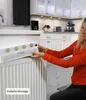A woman is installing a white heating amplifier on a radiator in a kitchen. Produktbild-Vorschau