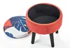 A round ottoman with a removable lid, shown on a white background. The ottoman is orange and has black legs. The lid has a blue monstera leaf pattern on a background of orange and white. Podgląd zdjęcia produktu