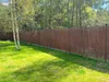 A brown reed fence is installed in a backyard, bordering a wooded area. The fence is surrounded by green grass. Produktbild-Vorschau