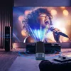 A projector is set up in a living room, projecting an image of a singer onto a large screen. Sound waves are visualized emanating from speakers and the projector. Náhľad obrázku produktu