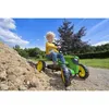 A young boy is riding a green and yellow pedal tractor in an outdoor, sandy area with trees and a blue sky in the background. Podgląd zdjęcia produktu