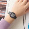 A person's wrist wearing a digital watch, with a book or magazine in the background. Anteprima dell’immagine prodotto