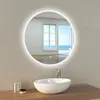 A round LED bathroom mirror with a light ring is mounted on a wall above a white vessel sink. The mirror reflects a room with large windows opening to a sunny ocean view. A faucet is visible above the sink, and a small box and jar are on the wooden counter Produktbild-Vorschau
