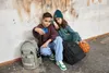 Two children are sitting on the ground next to backpacks, with a basketball nearby. The boy is holding up a phone to take a selfie. Produktbild-Vorschau