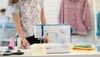 A woman is interacting with a sewing machine on a table, with various sewing supplies around. Produktbild-Vorschau