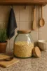 A glass jar filled with oats sits on a countertop next to a wooden cutting board and a bowl. Wooden spoons and a dark towel hang on hooks above the counter. Produktbild-Vorschau