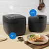 Two black rice cookers of different sizes are shown on a kitchen counter, with a plate of salmon and broccoli in the foreground. Produktbild-Vorschau