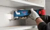 A person wearing gloves is using a Bosch multi-cutter to cut a square hole in a white cabinet. Podgląd zdjęcia produktu