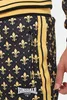 Close-up of a person's leg wearing black track pants with a gold fleur-de-lis pattern and gold stripes. Náhled obrázku produktu