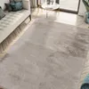 A plush beige rug is laid out on a wooden floor in a living room setting, with a sofa and coffee table visible. Produktbild-Vorschau