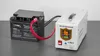 A black battery is connected to a white inverter on a gray surface. Produktbild-Vorschau