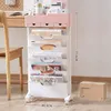 A mobile storage rack with multiple drawers and shelves, filled with books and magazines, is placed next to a wooden desk and chair. Produktbild-Vorschau