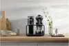 A black coffee maker is displayed on a wooden countertop with kitchenware and a vase with greenery. Produktbild-Vorschau