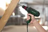 A hand is holding a green and black corded drill and drilling into a wooden plank. The background is blurred, suggesting an indoor workshop or construction site. Produktbild-Vorschau