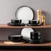 A set of stoneware dishes is displayed on two wooden shelves against a gray background. The dishes are black with a speckled white interior and a speckled gray rim. Two wooden salt and pepper grinders are on the top shelf. Produktbild-Vorschau