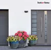 Three flower pots with colorful flowers are placed in front of a modern house entrance with a garage door and a front door. Náhled obrázku produktu