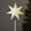 A white star-shaped tree topper is shown against a grey wooden background, with a blurred Christmas tree and lights to the right. Podgląd zdjęcia produktu