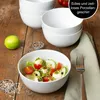 A close-up shot of a salad in a white bowl, with a stack of white bowls in the background, a lime, and spices on a wooden table. Produktbild-Vorschau