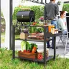 A grill cart with food and drinks is set up outdoors in a garden setting, with two people in the background. Produktbild-Vorschau