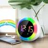 A digital alarm clock with a colorful LED ring is displayed on a wooden surface, next to a smartphone and earbuds. A framed picture with a rainbow and starfish is in the background, along with a glass vase containing a plant. Produktbild-Vorschau
