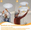 Two men are shown installing a ceiling light fixture. The first man is attaching the base to the ceiling, and the second man is attaching the light fixture to the base. Produktbild-Vorschau