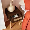A dark wood nightstand with a rattan drawer front sits next to a bed with orange bedding. A vase with dried flowers is on top of the nightstand, and a book is in the open drawer. Produktbild-Vorschau