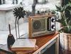 A vintage-style radio sits on a wooden table next to a decorative flower and some books, with a plant in the background. Podgląd zdjęcia produktu