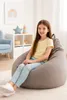 A young girl is sitting on a large, gray beanbag chair in a room with a light-colored wall and a blue armchair in the background. There is a framed picture of a rainbow on the wall and some children's toys on a shelf. Produktbild-Vorschau