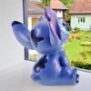 A blue Stitch figurine is sitting on a windowsill, looking out at a garden with houses. Produktbild-Vorschau