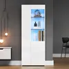 A modern white display cabinet with illuminated shelves, showcasing decorative items and books. A minimalist side cabinet is visible to the left, and a chair is in the background. Produktbild-Vorschau